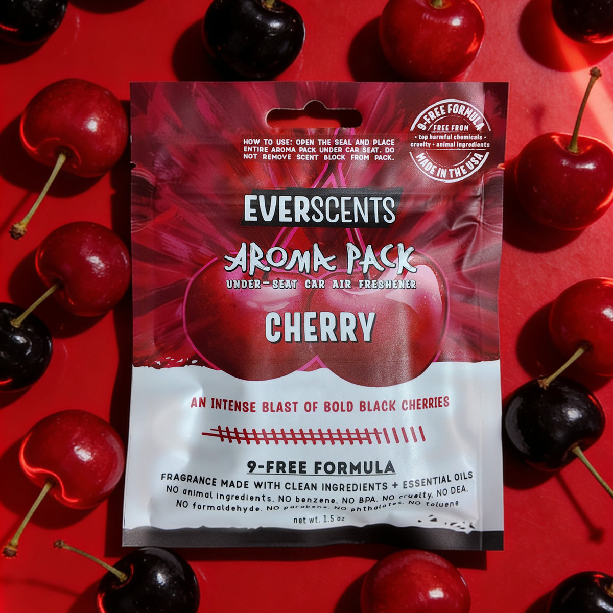 A flat-lay scene of an Everscents Aroma Pack under-seat car air freshener pouch in the Cherry scent lying flat and artfully surrounded by fresh glossy red and black cherries on a vibrant red studio background. The scene evokes a fun and fruity atmosphere.