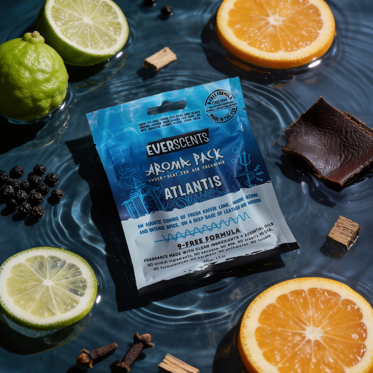 A flat-lay scene of an Everscents Aroma Pack under-seat car air freshener pouch in the Atlantis scent styled on a deep muted blue watery background surrounded by an artful scattering of kaffir lime slices, orange slices, peppercorns, cloves, leather, and driftwood pieces. The scene is dramatically dimly-lit, evoking a masculine high-end luxury aesthetic.