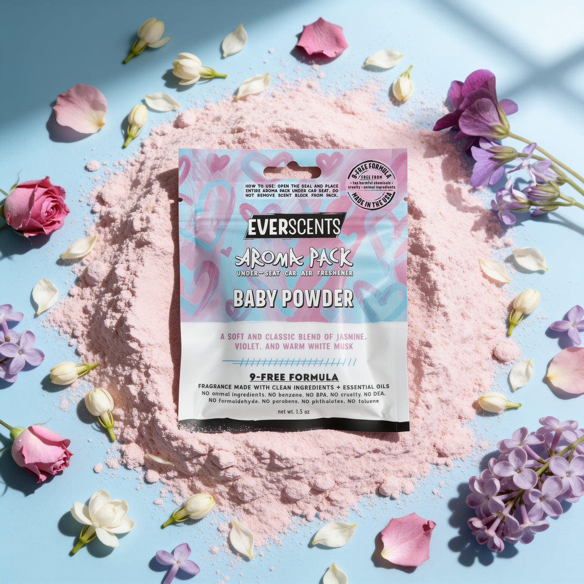 A flat-lay scene of an Everscents Aroma Pack under-seat car air freshener pouch in the Baby Powder scent styled on a pile of pale pink powder surrounded by an elegant scattering of small white flower petals, lilac, violet, and roses on a matte baby blue studio background surface. The scene is lit by a soft overhead light casting shadows of a nearby windowpane onto the surface. The scene evokes a high-end graceful clean  aesthetic.
