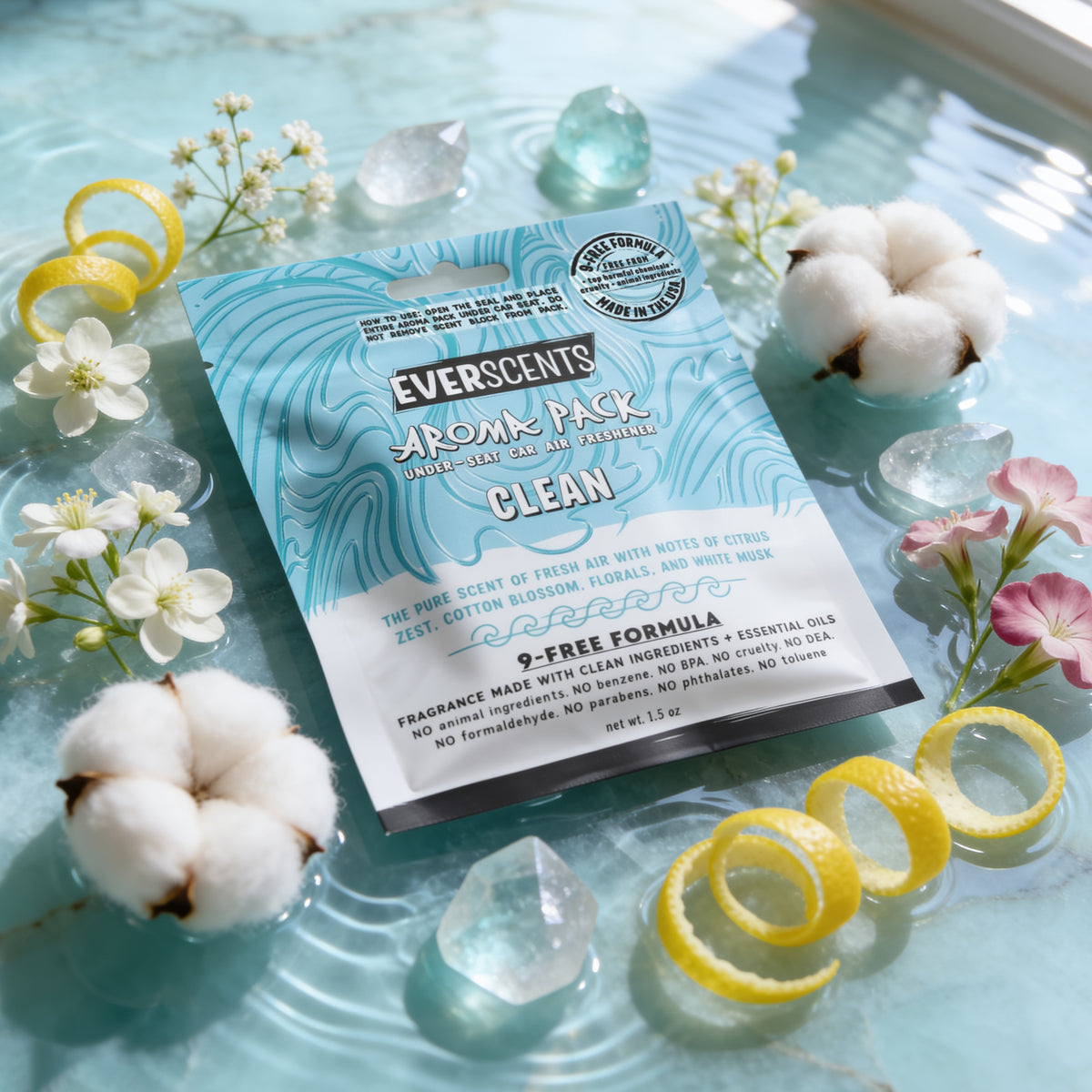 A flat-lay scene of an Everscents Aroma Pack under-seat car air freshener pouch in the Clean scent styled on a light blue quartz stone slab covered in shallow lightly rippling water and surrounded by an elegant scattering of cotton blossoms, lemon zest curls, small flowers, and quartz crystals. The scene is serene and brightly-lit, evoking a clean, high-end luxury aesthetic.