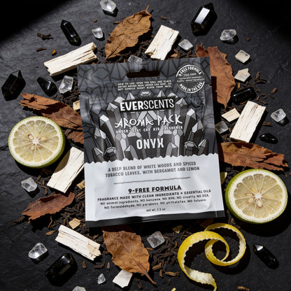 A flat-lay scene of an Everscents Aroma Pack under-seat air freshener pouch in the Onyx scent styled on a slate stone background surrounded by a dynamic scattering of dried tobacco leaves, sliced bergamot, lemon zest curls, white wood pieces, and small onyx and quartz crystals. The scene is dramatically-lit by an overhead studio light, evoking a moody luxury atmosphere with a masculine aesthetic.